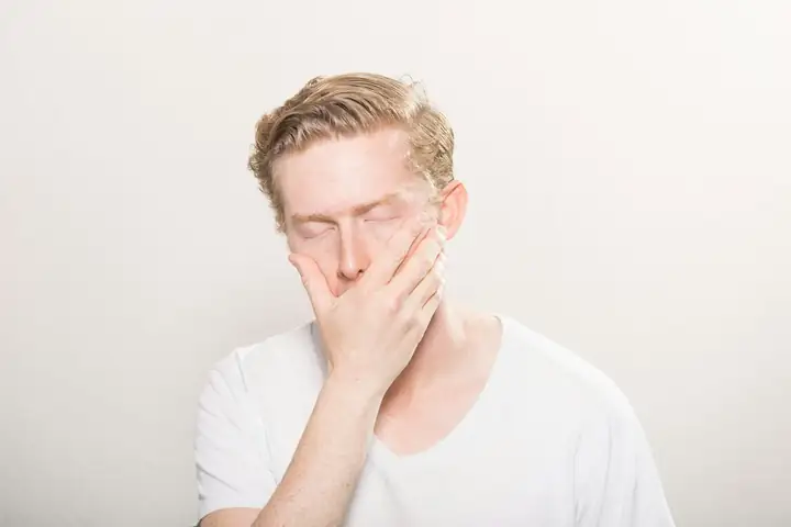 Man in a white T-shirt covering his nose and mouth with one hand, eyes closed