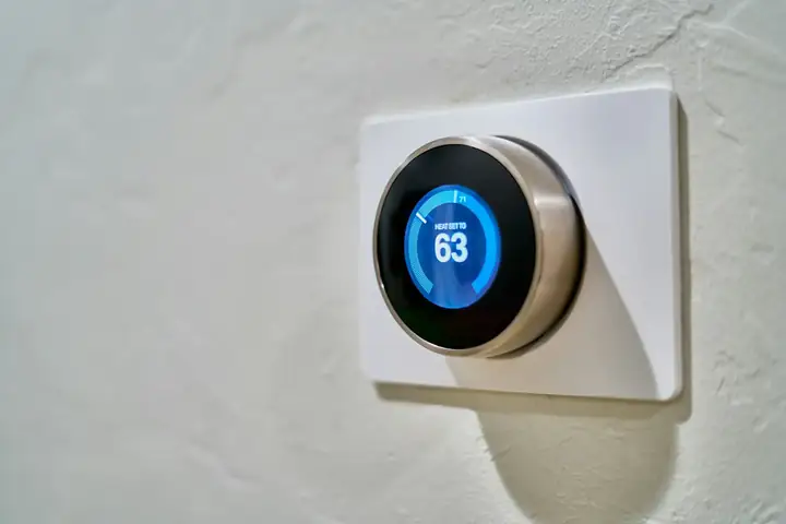Wall-mounted smart thermostat with a blue display reading heat set to 63, seen at an angle